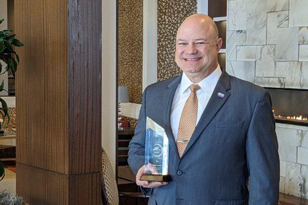 Greg Accardo hold an award. He is wearing a grey suit. 