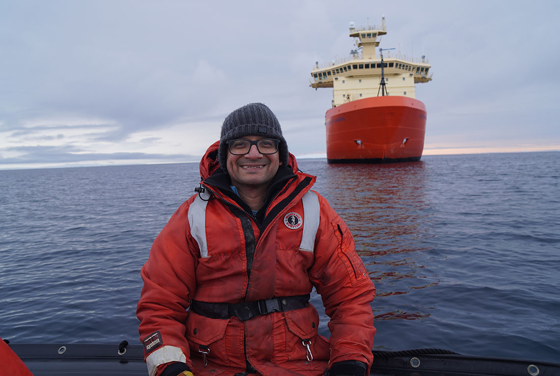 A man wearing an orange parka sits on a boat with a ship in the background