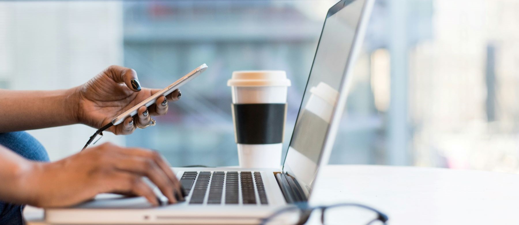 closeup of two hands; one holds a phone, and the other types on a laptop